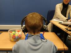 A child wearing headphones sits at a table with colorful plastic pieces in a container in front. In the background, an adult is seated at another table, working with papers and equipment. 