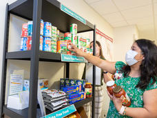 A woman wearing a surgical mask places jars and cans of food on a shelf of a food pantry